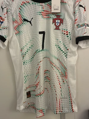 Portugal fotbollströja Ronaldo #7 Puma - Portugal landslagströja från Puma med nummer 7 och Ronaldo på ryggen. Tröjan är vit med mönster i grönt och rött, svarta detaljer och landslagets emblem på bröstet. Korta ärmar och lätt, ventilerande material perfekt för fotbollsträning eller match. Helt oanvänd, kommer med etikett. 