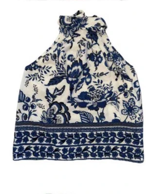 Blommig halterneck linnetopp - Supersöt halterneck topp med blått blommönster på vit botten. Toppen har hög hals med knyt och är ärmlös, perfekt för varma dagar. Lätt och luftig känsla, passar till jeans eller kjol.