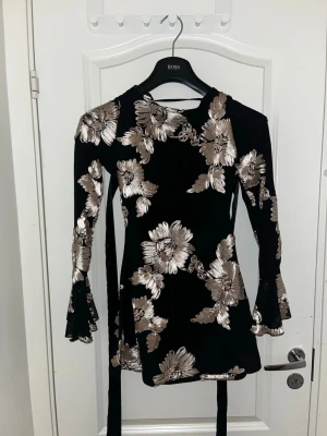 Svart klänning med guldblommor - Supersnygg svart klänning med stora guldiga blommor och trumpetärmar i mesh. Klänningen har figurnära passform och markerad midja. Perfekt för dig som vill sticka ut med ett unikt mönster och trendiga detaljer.