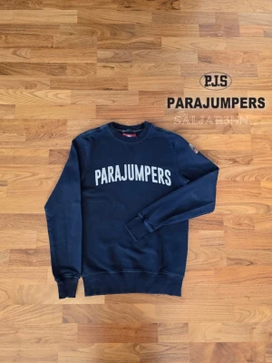 Parajumpers Sweatshirt – Marinblå | Storlek S - Parajumpers Sweatshirt – Marinblå | Storlek S Stilren Parajumpers sweatshirt i marinblå med stor vit logotyp framtill som ger en sportig och modern look. Tillverkad i mjuk bomullsfleece som håller dig varm och bekväm. Lång ärm med ribbade fållar och ärmslut ger bra passform. Märkespatch på ärmen och QR-kod för äkthetskontroll finns kvar. Plagget är i nyskick. 📦 Snabb leverans – Kontakta för frågor!