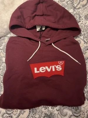 Vinröd hoodie från Levi's - Snygg vinröd hoodie från Levi's med klassisk röd logga framtill. Hoodien har vita dragsnören, stor huva och en magficka. Tillverkad i mjukt bomullsmaterial och har långa ärmar med muddar. Perfekt för en avslappnad och trendig stil.