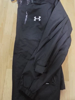 Svart vindjacka från Under Armour - Svart vindjacka från Under Armour med huva och dragkedja. Jackan har vit logotyp på bröstet, elastiska muddar och diskreta detaljer. Materialet är lätt och vattenavvisande, perfekt för blåsiga dagar. Snygg och sportig design med praktiska fickor.