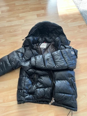 Moncler maya M - Snygg svart dunjacka från Moncler med glansig finish och huva. Jackan har dragkedja framtill, två fickor med dragkedja och Moncler-logga på ärmen. Perfekt för kalla vinterdagar och riktigt trendig stil.
