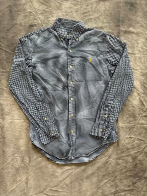 Blårutig skjorta från Polo Ralph Lauren - Säljer en blårutig skjorta från Polo Ralph Lauren med slim fit-passning. Skjortan har klassisk krage, knappar framtill och det ikoniska gula Polo-logot broderat på bröstet. Perfekt för dig som gillar stilrena och tidlösa plagg.