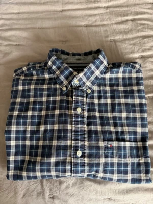 Blårutig skjorta från Tommy Hilfiger - Snygg blårutig skjorta från Tommy Hilfiger med klassisk krage och knappar framtill. Skjortan har lång ärm och en liten logga på bröstfickan. Perfekt för dig som gillar en stilren och tidlös look.lite sliten  längst up vid halsen 