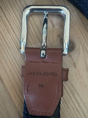 Blått flätat bälte från Jack & Jones - Snyggt blått flätat bälte från Jack & Jones med detaljer i brunt skinn och ett silvrigt metallspänne. Perfekt accessoar för att lyfta din outfit och funkar till både jeans och chinos. Längd 80 cm enligt märkning.