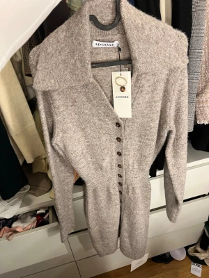 Beige stickad klänning från Adoore - Mysig grå stickad klänning från Adoore med markerad midja och långa ärmar. Klänningen har en djup v-ringning och knappar framtill hela vägen ner. Perfekt för dig som gillar en enkel men stilren look. Storlek 32 men passar mig med 34 då den är väligt stretchiga 