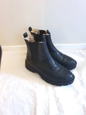 Svarta fodrade boots ifrån nelly - Säljer ett par svarta boots med chunky sula och elastiska paneler på sidorna. Insidan är fodrad med mjukt, ljust fuskpälsfoder som håller dig varm. Klassisk dragflik både fram och bak för enkel påtagning. Perfekta för kyliga dagar.