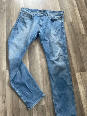 Replay Anbass Jeans - Säljer ett par välanvända och sköna jeans från kvalitetsmärket Replay, modell "Anbass". Jeansen har en snygg ljusblå tvätt med naturligt slitage som ger en avslappnad look. De har klassisk 5-ficksdesign och den ikoniska Replay-loggan på baksidan av linningen och lilla flaggan vid framfickan. Passformen är Anbass, vilket brukar betyda en smal men bekväm passform (slim) 