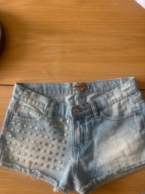 Ljusblå jeansshorts med nitar - Låg midjade Snygga ljusblå jeansshorts från Lemonissima med slitna detaljer och råa kanter. Ena framsidan är dekorerad med silvriga nitar för en cool look. Klassisk femficksmodell och knappgylf. Perfekta för sommaren!