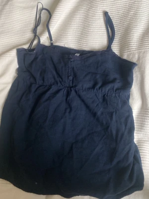 Mörkblå linnetopp med spetsdetaljer - Söt mörkblå linnetopp med smala axelband och spetsdetaljer längs urringningen. Toppen har dekorativa knappar framtill och är tillverkad i ett mjukt material som känns skönt mot huden. Perfekt för varma dagar eller som lager under en kofta.