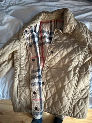 Beige quiltad jacka från Burberry - Klassisk beige quiltad jacka från Burberry med det ikoniska rutiga fodret. Jackan har knappar framtill, krage och lång ärm. Perfekt för dig som gillar tidlös och stilren design.