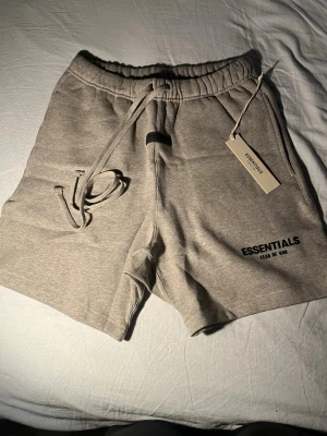 Dark oatmeal Essentials shorts med snörning - Snygga dark oatmeal shorts från Essentials Fear of God med snörning i midjan och elastisk linning. Mjuk bomullskvalitet och diskret logga på benet. Perfekta för en avslappnad och trendig look.