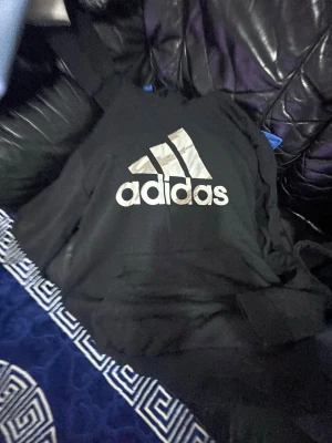 Svart hoodie från Adidas med logga - Svart hoodie från Adidas med stor vit logga på bröstet. Klassisk modell med huva och magficka. Tillverkad i mjukt material som passar perfekt för en avslappnad streetwear-look. Ribbade muddar vid ärmar och nederkant.
