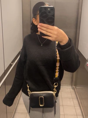 Svart handväska från Marc Jacobs - Cool svart handväska från Marc Jacobs med guldiga detaljer och bred axelrem med text. Väskan har en rektangulär form och är tillverkad i läder. Perfekt för dig som vill ha en trendig och stilren accessoar. Om ni söker så kan ni se att de kostar från 4000-3000 så jag tänker sälja för 2500 och om ni är seriösa och köper direkt så kan vi lätt prata om priset 