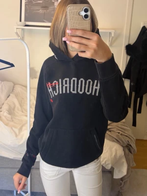Svart hoodie från Hoodrich - Cool svart hoodie från Hoodrich i en med stor logga på bröstet och detaljer i rött. Har huva och känguruficka framtill. Texttryck på ryggen/vänster handled och broderade detaljer på huvan. Perfekt för dig som gillar streetwear och vill ha något unikt!