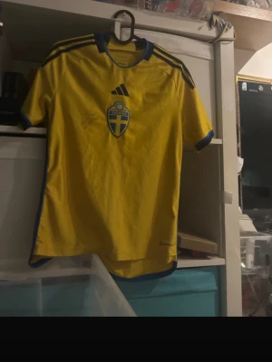 Svensk landslagströja Adidas gul - Gul svensk landslagströja från Adidas med blå detaljer och Sveriges emblem på bröstet. Tröjan har korta ärmar, rund hals och texten 'Sverige' på ryggen. Perfekt för fotbollsfans som vill visa sitt stöd. Materialet är lätt och andas, idealiskt för sport.