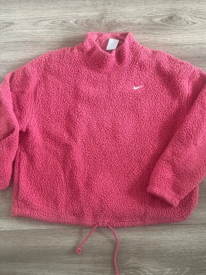 Rosa fleece-tröja från Nike - Mysig rosa fleece-tröja från Nike med Therma-FIT-teknologi. Tröjan har hög krage, dragsko nertill och broderad vit Nike-logga på bröstet. Perfekt för kyliga dagar och enkel att styla till jeans eller leggings.