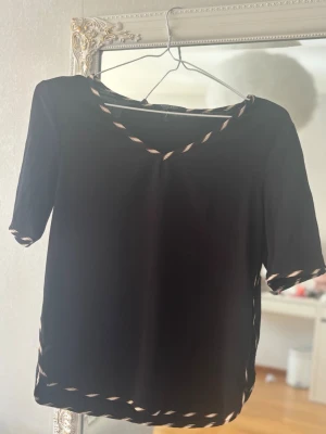 Svart topp från Scotch & Soda - Svart topp från Scotch & Soda med korta ärmar och dekorativa band i beige och brunt längs halsringning, ärmslut och nederkant. Toppen har en avslappnad passform och är perfekt för en trendig och stilren look.