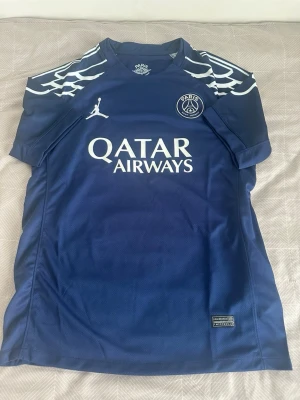 PSG matchtröja blå Jordan - Säljer en blå Paris Saint-Germain matchtröja från Jordan med vita detaljer och vingmönster på axlarna. Tröjan har PSG-logga, Jordan-logga och Qatar Airways-tryck på bröstet. Perfekt för fotbollsträning eller att bära som supporter.