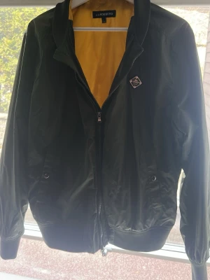 J.Lindeberg jacka - Stilren svart bomberjacka från J.Lindeberg med gul insida och klassisk ribbad krage. Jackan har dragkedja framtill, två sidofickor med knapp och en snygg logotyp-patch på bröstet. Materialet är glansigt och lätt, perfekt för en trendig streetwear-look. Storlek L passar M