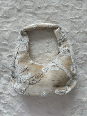 Beige axelväska med broderade blommor - Trendig beige handväska i mjukt tyg med broderade blommor i vitt och grått. Väskan har en rundad form och bred axelrem, perfekt för att bära över axeln. Unik design med tydliga blommönster som ger en cool och personlig stil.