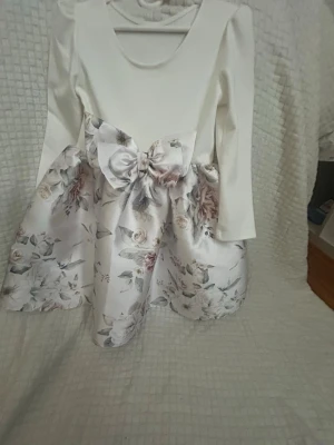 Vit blommig klänning med rosett - Söt vit klänning med lång ärm och stor rosett i midjan. Kjolen har ett romantiskt blommönster i ljusa toner och tyget känns glansigt och elegant. Perfekt för speciella tillfällen och ger en klassisk look. Vet ej storlek borde passa mellan 5-7år 
