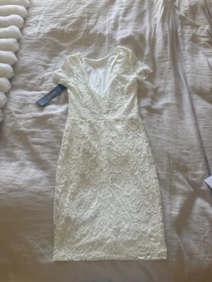 Vit spetsklänning med v-ringning - Säljer en vit spetsklänning med v-ringad hals och korta ärmar. Klänningen har ett blommigt spetsmönster och är perfekt för konfa/skolavslutning/student