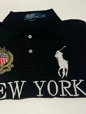 Svart pikéskjorta från Ralph Lauren - Svart pikéskjorta från Ralph Lauren med stora broderade detaljer, bland annat en vit polospelare och texten 'NEW YORK' framtill. Skjortan har krage, knappar och ett broderat emblem i guld och rött på bröstet.