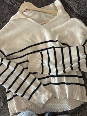Beige stickad tröja med svarta ränder - Snygg beige stickad tröja med svarta horisontella ränder. Tröjan har v-ringning, långa ärmar och ribbade muddar vid ärmslut och nederkant. Perfekt för en avslappnad och trendig look. Supersnygg med en piké eller skjorta under!