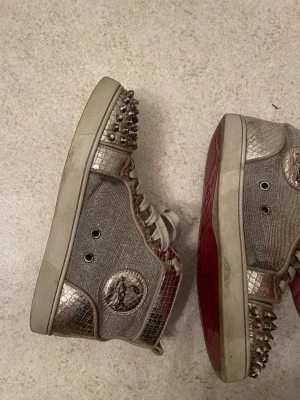 Metallic high top sneakers med nitar - Unika high top sneakers i metallic silver med ormskinnsmönster, dekorerade med silvriga nitar på tå och häl. Skorna har vita snören, röd sula och ett rundat märke på sidan. Perfekta för dig som vill sticka ut med din stil.