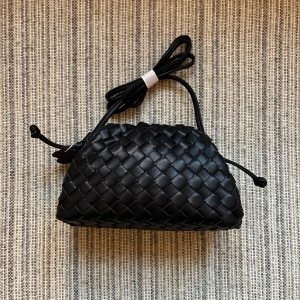 Svart flätad handväska - Snygg svart handväska med justerbar axelrem. Väskan har en mjuk, rundad form och stängs med dragsko i sidorna. Perfekt för dig som gillar stilrena accessoarer med en twist.