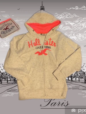 Beige hoodie från Hollister med dragkedja - Snygg beige hoodie från Hollister med dragkedja och huva. Stor röd logga och texten 'Hollister California' framtill. Insidan av huvan är neonröd för en cool kontrast. Mjuk bomullsblandning och två fickor framtill. Perfekt för en avslappnad streetstil.strl 164