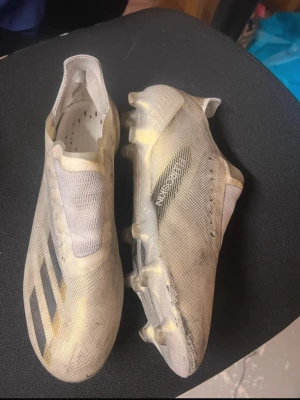 Vita Adidas X Ghosted+ fotbollsskor - Säljer ett par vita Adidas X Ghosted+ fotbollsskor med svarta detaljer och klassiska tre ränder på sidan. Skorna har en strömlinjeformad design utan snörning och ovandel i lätt syntetmaterial för maximal fart på planen. Perfekta för dig som vill ha speed och stil på matchen.