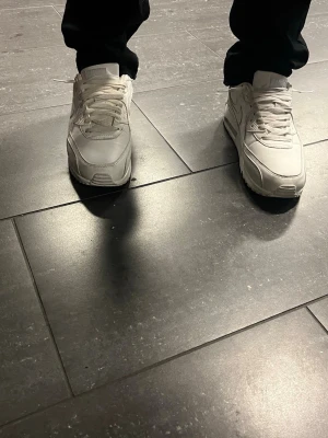 Vita Nike Air Max 90 sneakers - Säljer ett par klassiska vita Nike Air Max 90 sneakers i skön läderlook. De har den ikoniska swoosh-loggan på sidan, tjock sula med synlig Air-enhet och vita skosnören. Perfekta för dig som gillar stilrena och tidlösa sneakers.