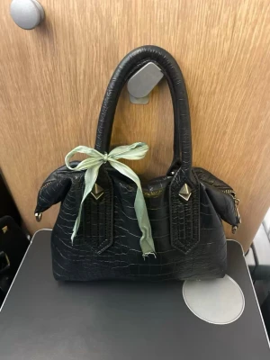 Svart handväska med krokopräglat mönster - Snygg svart handväska i krokopräglat konstläder med två handtag och guldfärgade detaljer. Väskan har en ljusgrön rosett knuten på handtaget för en personlig touch. Perfekt storlek för det viktigaste och ger en cool vibe till din outfit. Den är lite sliten vid handtagen då skinnet har gått av, varav är priset diskuterbart!