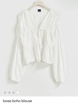 Vit boho-blus med knytband - Luftig vit boho-blus med v-ringning och knytband framtill. Blusen har långa ärmar med lätt puff och är tillverkad i ett tunt, strukturerat tyg som ger en avslappnad känsla. Perfekt att styla med jeans för en trendig look.