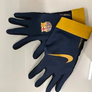 Nike FC Barcelona handskar - Snygga blå handskar från Nike med FC Barcelona-logga och gula detaljer. Handskarna har svart insida, gul mudd och är perfekta för dig som vill visa ditt lagstöd även när det är kallt. Materialet är syntet och designen är sportig med Nike Swoosh och klubbmärke.