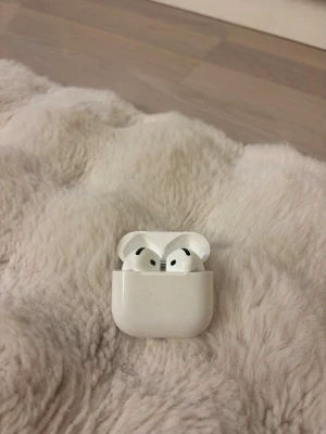Airpods 4 trådlösa hörlurar - Säljer ett par vita Apple AirPods med matchande laddningsetui. Hörlurarna är trådlösa och har en stilren, rundad design i blank plast. Perfekta för musik, samtal och poddar när du är på språng.