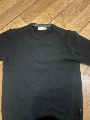 Svart stickad tröja från - Klassisk svart stickad tröja från  med rund halsringning och ribbade muddar. Enkel och stilren design som passar till det mesta. Perfekt basplagg för garderoben.