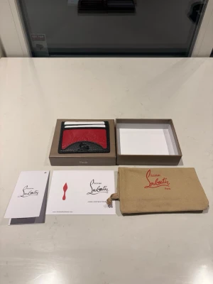  röd kortfodral - Säljer ett exklusivt kortfodral i svart och rött läder. Fodralet har plats för flera kort och pryds av den ikoniska loggan i relief på framsidan. Kommer med originalbox, dustbag och äkthetskort.