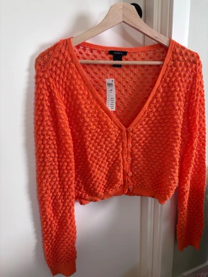 Orange virkad kofta från Lindex - Trendig orange virkad kofta från Lindex med v-ringning och knappar framtill. Koftan har lång ärm och kortare, boxig passform. Perfekt för lager på lager och ger en cool touch till din outfit.