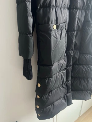 Svart lång dunjacka från Busnel - Snygg svart lång dunjacka från Busnel med guldfärgade knappar och diskret B-logga. Jackan har stora fickor framtill och ribbade muddar vid ärmsluten. Perfekt för kalla vinterdagar och ger en stilren look.