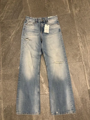Ljusblå raka jeans med slitningar - Säljer ett par ljusblå jeans med raka ben och slitningar vid knäna. Jeansen har en avslappnad passform, klassiska fem fickor och är tillverkade i denim. Perfekta för en cool och trendig look. Helt oanvända 