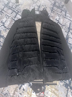 Svart jacka från Canada Goose - Snygg svart dunjacka från canada goose. jackan har horisontella sömmar, två dragkedjeförsedda fickor och hög krage. Perfekt för lager-på-lager och håller dig varm under kyliga dagar. OBS det är en kopia av en riktig canada goose men ser ut som en äkta och helt oanvänd med etiketten på
