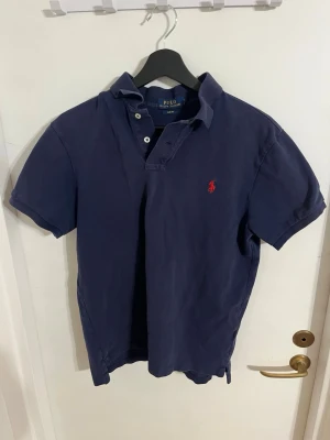 Marinblå pikétröja från Polo Ralph Lauren - Klassisk marinblå pikétröja från Polo Ralph Lauren med korta ärmar och krage. Tröjan har två knappar vid halsen och den ikoniska röda logotypen broderad på bröstet. Perfekt till jeans eller shorts för en avslappnad men stilren look.