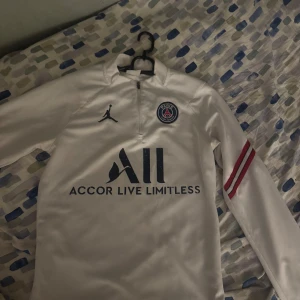 Vit PSG långärmad fotbollströja Nike - Snygg vit långärmad PSG-tröja från Nike med dragkedja vid halsen. Tröjan har Paris Saint-Germain-logga, Jordan-logga och Accor Live Limitless-tryck på bröstet samt två röda ränder på ena ärmen. Perfekt för dig som älskar fotboll och vill sticka ut.