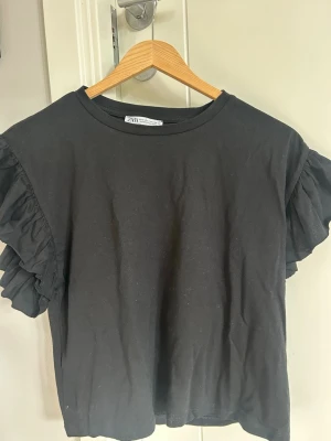 Svart volangärmad topp från Zara - Säljer en svart topp från Zara med volangdetaljer på ärmarna. Toppen har rund halsringning och är gjord i mjuk bomull. Perfekt för dig som vill ha en enkel men ändå trendig look.