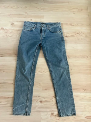 Blå raka jeans från Nudie Jeans - Klassiska blå jeans från Nudie Jeans med rak passform och fem fickor. Jeansen har Nudie Jeans signaturbroderi på bakfickorna och en brun patch i midjan. Tillverkade i denim med en snygg tvättad look. Vältvättade men väldigt fina