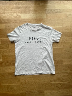 Vit t-shirt från Polo Ralph Lauren - Klassisk vit t-shirt från Polo Ralph Lauren med svart logotyptryck framtill. T-shirten har rund halsringning och korta ärmar. Tillverkad i mjuk bomull för en skön och avslappnad stil.
